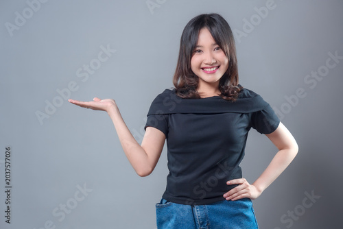 A cheerful Asian woman wearing a black top and denim skirt smiling confidently while pointing with one hand, showing a welcoming and presenting gesture suitable for advertising or promotional concept.
