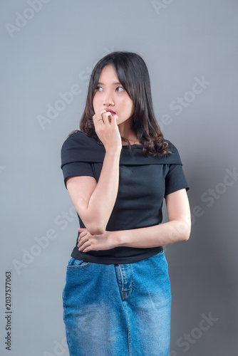 An Asian woman wearing a black top and denim skirt looks thoughtful and curious while touching her lips, showing a pensive expression that reflects contemplation and inner emotion.