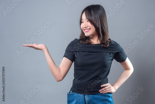 A smiling Asian woman wearing a black top and denim skirt gestures with her hand to the side, showing a confident and friendly expression, perfect for presentation or advertising concepts.