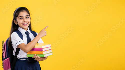 Happy Indian schoolgirl holding books pointing at yellow copy space
