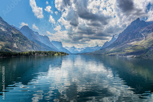 Turquoise Saint Mary Lake surrounded by steep forested mountain slopes and weathered tree stumps under a bright blue Montana sky in Glacier National Park