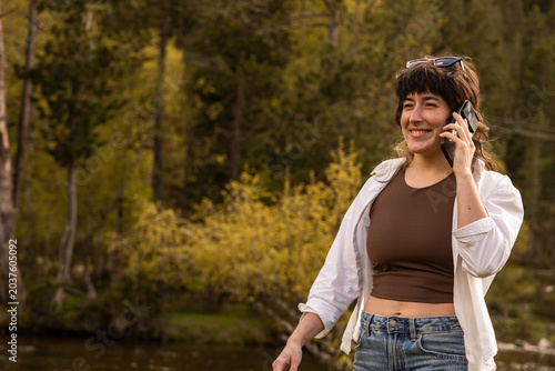 Woman wears a white jacket and brown top. She holds a phone to her ear, smiling. Sunglasses rest on her head. Trees and a lake surround her. The scene feels peaceful