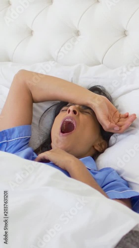 Vertical video: Yawning woman in blue pjs waking, stretching, settling in bed to rest with duvet