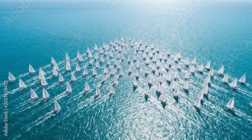 Aerial view of a summer regatta with numerous sailboats racing in formation across the sparkling blue ocean under clear skies, capturing vibrant energy and harmony