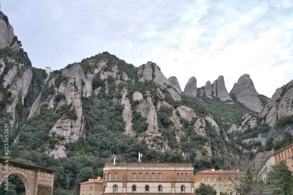 Fototapeta premium The unique jagged peaks of Montserrat mountain range rise majestically above lush green vegetation and buildings below