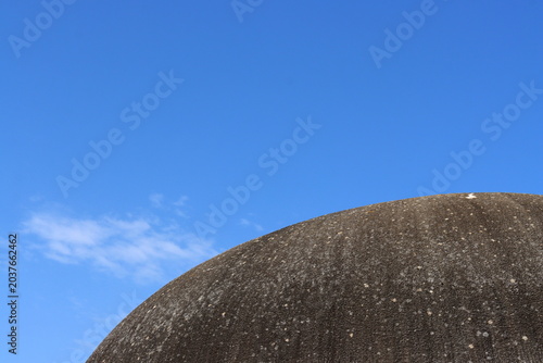 Round textured roof surface against a clear blue sky with soft white clouds, creating a minimal architectural composition with strong contrast between structure and open space.