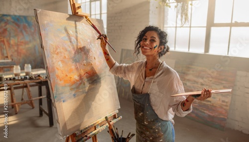 Joyful woman artist smiling while holding paintbrushes and painting colorful abstract artwork in bright studio with natural light