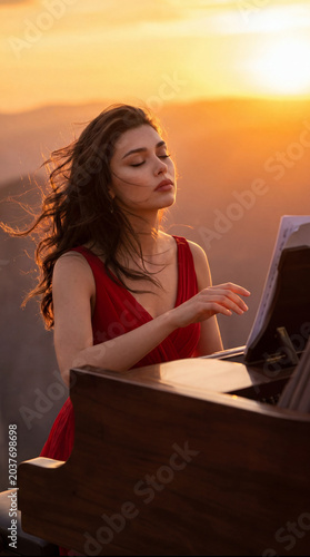 Musician plays piano at sunset on a mountain with long hair blowing in the wind while wearing a red dress