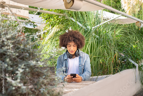 Young mixed race boy ,dressed in casual clothes using smartphone in green area in nature. Green open space for coworking, freelance, relax, coffee, food. Free place to work, business, study, freelance