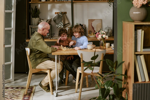Senior Caucasian man playing chess with two kids at home, teaching strategy and bonding across generations. Useful for family lifestyle, parenting, retirement, education campaigns
