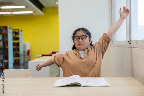 Tired young Asian girl student stretching arms at desk after reading book in classroom, sleepy child school kid relaxing from learning lesson inside library room
