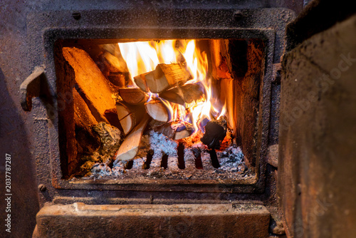 Close up of burning wooden logs blazing in fire flames inside wood burning stove with open glass door in cosy warm home living room.