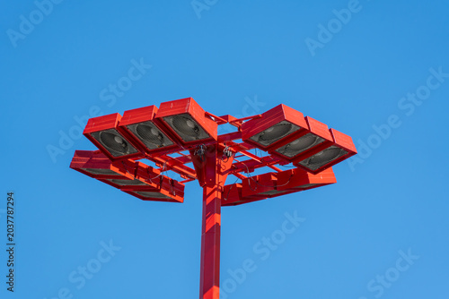 Red stadium floodlights fill the frame against a cloudless blue sky. The close angle shows metal housings, lenses, cables, and urban lighting detail.