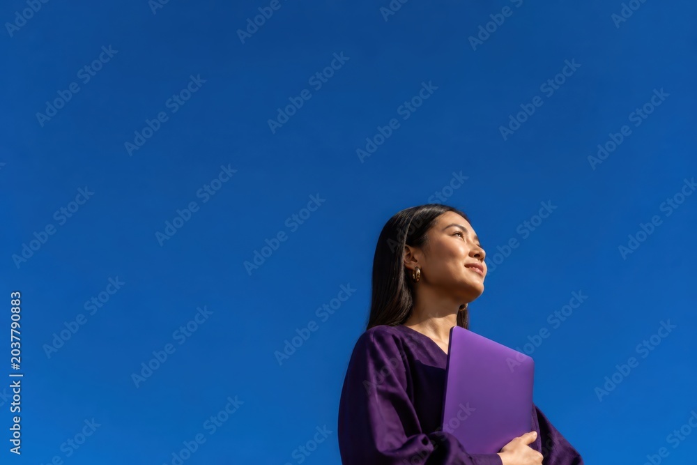 Obraz premium Confident young woman holding a purple folder against a clear blue sky in a calm outdoor portrait
