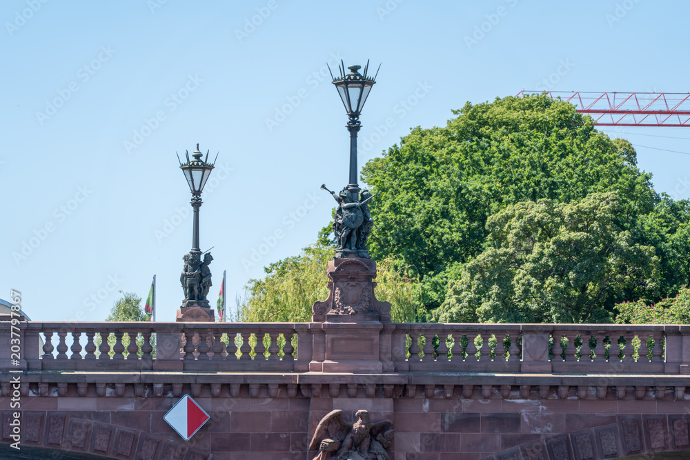 Obraz premium Decorative lanterns and sculpted figures top a stone bridge above the Spree in Berlin. Leafy deciduous trees and a crane frame the riverside landmark.