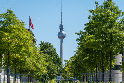 Schilderij op canvas The Berlin TV Tower stands between leafy deciduous trees with a red flag nearby