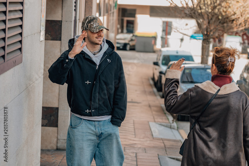 Young man in cap and jacket talking with a woman on an urban sidewalk, both gesturing expressively during a casual street conversation