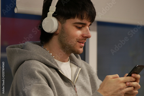 young man wearing white headphones and a gray hoodie smiles while using his phone in a bright, modern space