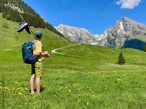 Barefoot boy hiking through rural Toggenburg, Switzerland, carrying a wooden stick over his shoulders with hiking shoes dangling from it.