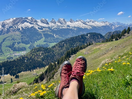 Feet wearing hiking boots resting in front of the panoramic view of the Churfirsten mountain range in the Swiss Alps, Toggenburg region, St. Gallen, Switzerland.