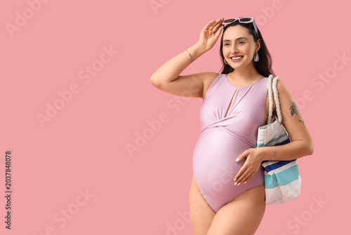 Young pregnant woman in swimsuit with handbag on pink background. Traveling c...
