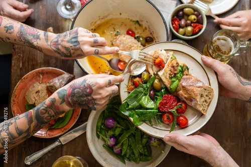Fototapeta na wymiar Tattooed hands sharing a meal with salad, bread, and olives on a table.