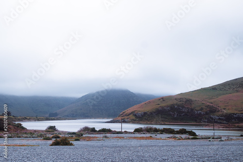 Photography Lake Forsyth, New Zealand: Calm water of Wairewa lagoon behind dark gravel stone ridge under moody low mist over rural hills, across Birdlings Flat