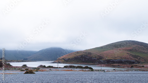 Photos Lake Forsyth, New Zealand: Wide panoramic view of calm Wairewa lagoon water with dark shingle gravel bank and dramatic white cloud fog over hills