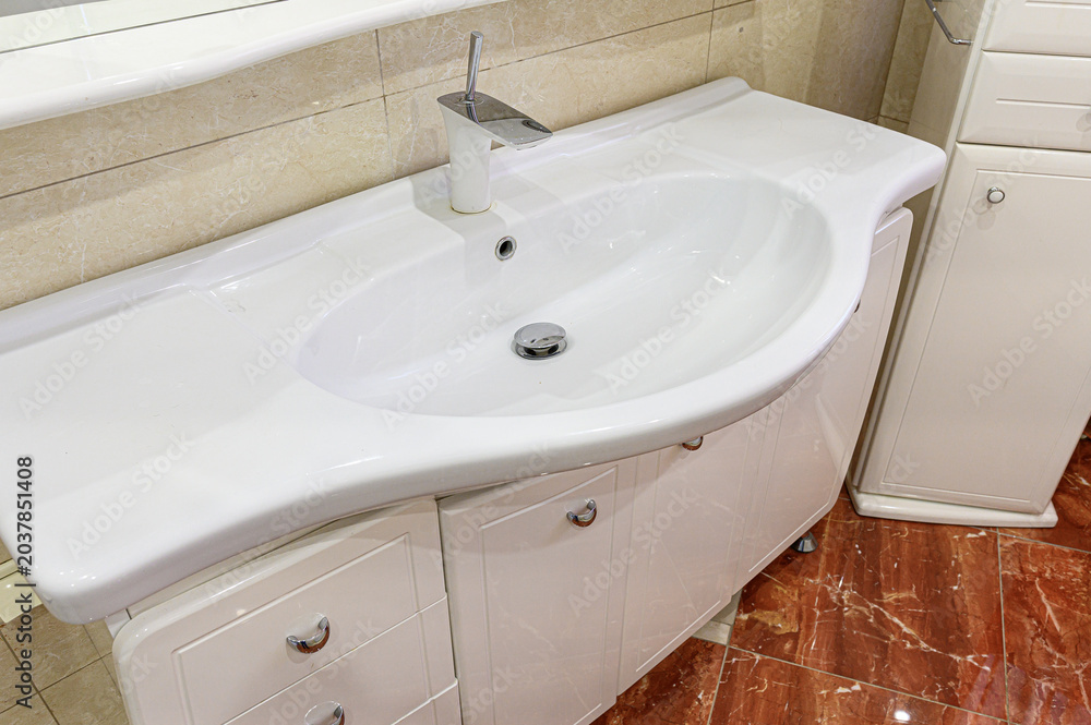 Fototapeta premium A white asymmetrical ceramic sink sits atop a glossy white vanity unit; paired with a chrome faucet, beige tiled walls, and a rich brown marble floor