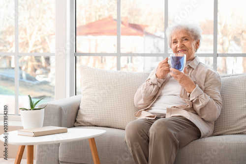 Tableau sur toile Senior woman with glass cup of butterfly pea flower tea sitting on sofa near win