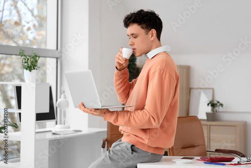Young man with laptop drinking coffee in office