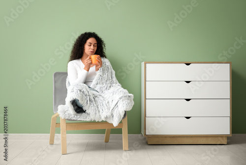 Young African-American woman with cup of tea and warm blanket sitting in whit...