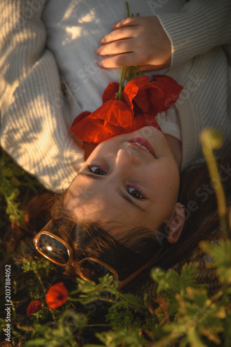 Portrait of little girl lying in blooming poppy field holding bouquet of red poppies and smelling flowers, warm childhood summer atmosphere and nature beauty concept