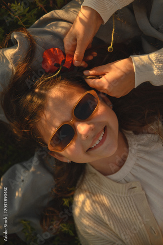 Two best friends relaxing in a blooming poppy field, one girl lying on the other’s lap while a red poppy flower is gently woven into her braid, capturing childhood friendship and warm summer moments
