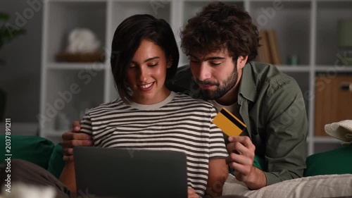 Couple enjoying online shopping with credit card and laptop at home while ordering goods from the couch at night