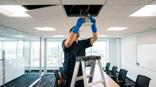 professional electrician on a stepladder installing and wiring an led recessed ceiling light fixture inside a corporate office conference boardroom