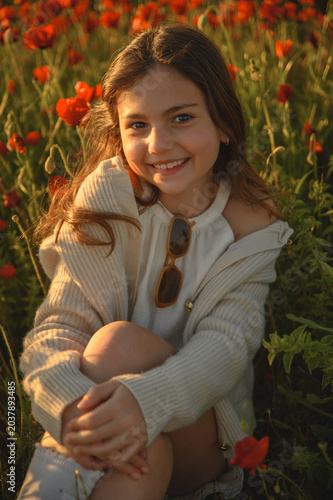 Little girl in light dress sitting in blooming poppy field at warm sunset light, smiling with joy, carefree summer childhood and nature lifestyle concept