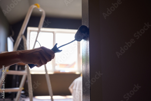 Foto Hand uses paint roller to apply dark paint along wall corner, with ladder in bac