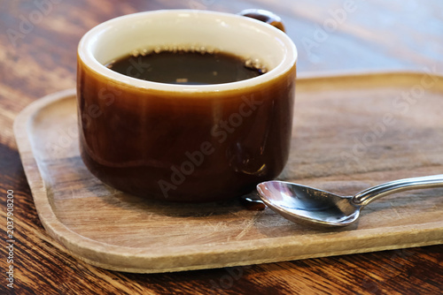 Black coffee in a brown ceramic cup on a wooden tray with a metal spoon, rustic tabletop .