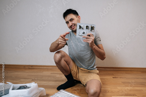 Canvas Print Smiling man holding blister pack of furniture parts and allen key, pointing finger towards camera, conveying sense of invitation, readiness, successful diy home improvement