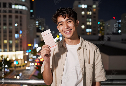 Smiling Person Holding Lottery Ticket in Front of City Night Lights