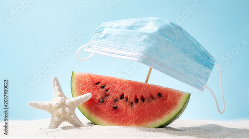 Watermelon slice arranged as a boat with medical mask sail on white sand