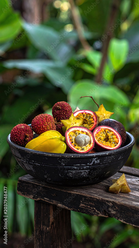 Obraz premium Vibrant bowl of exotic tropical fruits including juicy passion fruit, starfruit, and lychees on a rustic wooden bench against a lush green garden background. Perfect for healthy food concepts.