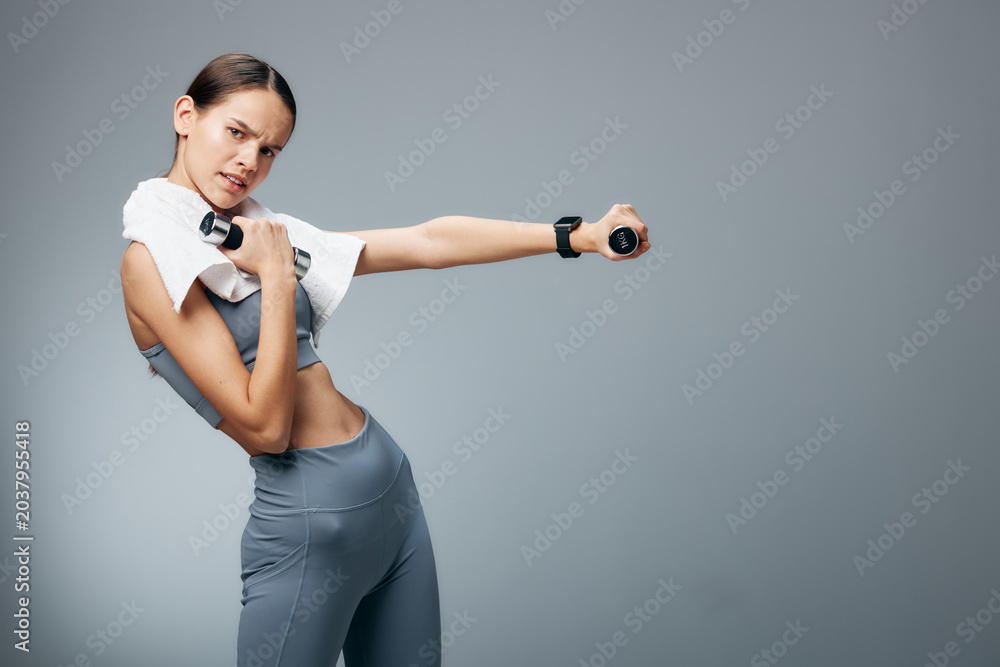 Fototapeta premium Athletic woman in workout gear stretches with dumbbells in a studio, towel on shoulder, showcasing fitness, strength training and confident movement.