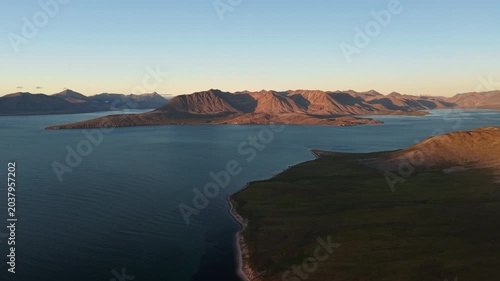 Drone cinematic shot pans over Gilmimyul Bay near Provideniya, Chukotka, Russia. Rugged mountains and calm sea glow in summer evening light. Smooth pan reveals remote Arctic coastline