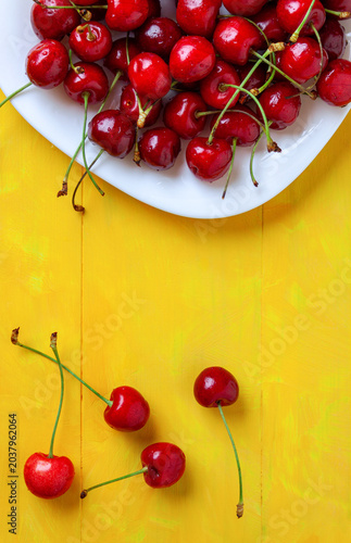 Top view of fresh red cherries on a white plate with scattered berries on a yellow wooden table. sweet summer fruit,