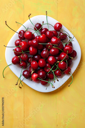 Top view of a white ceramic plate filled with ripe red cherries on a bright yellow wooden table.