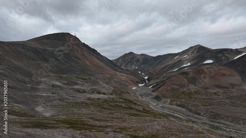 Wide mountain basin under heavy clouds in Russia, Chukotka, Provideniya. Aerial establishing shot slowly pans across tundra ridges, snow patches, scree fields and winding tracks in daylight.