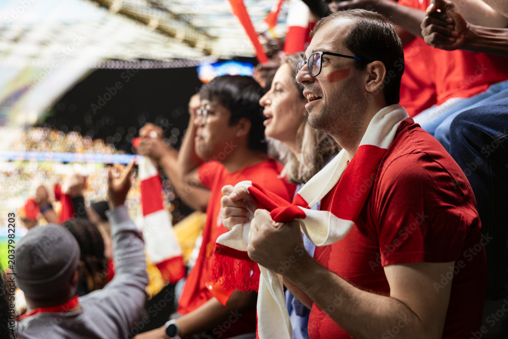 Obraz premium Anxious football supporters watching a decisive final attack during match from packed stadium stands with red scarves and fan tension. Supporter holding breath with nervous anticipation.