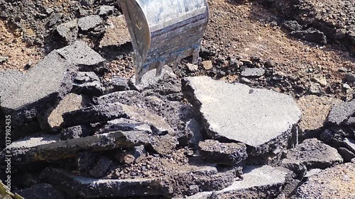 Excavator bucket pushing broken concrete slab piece across demolition site ground